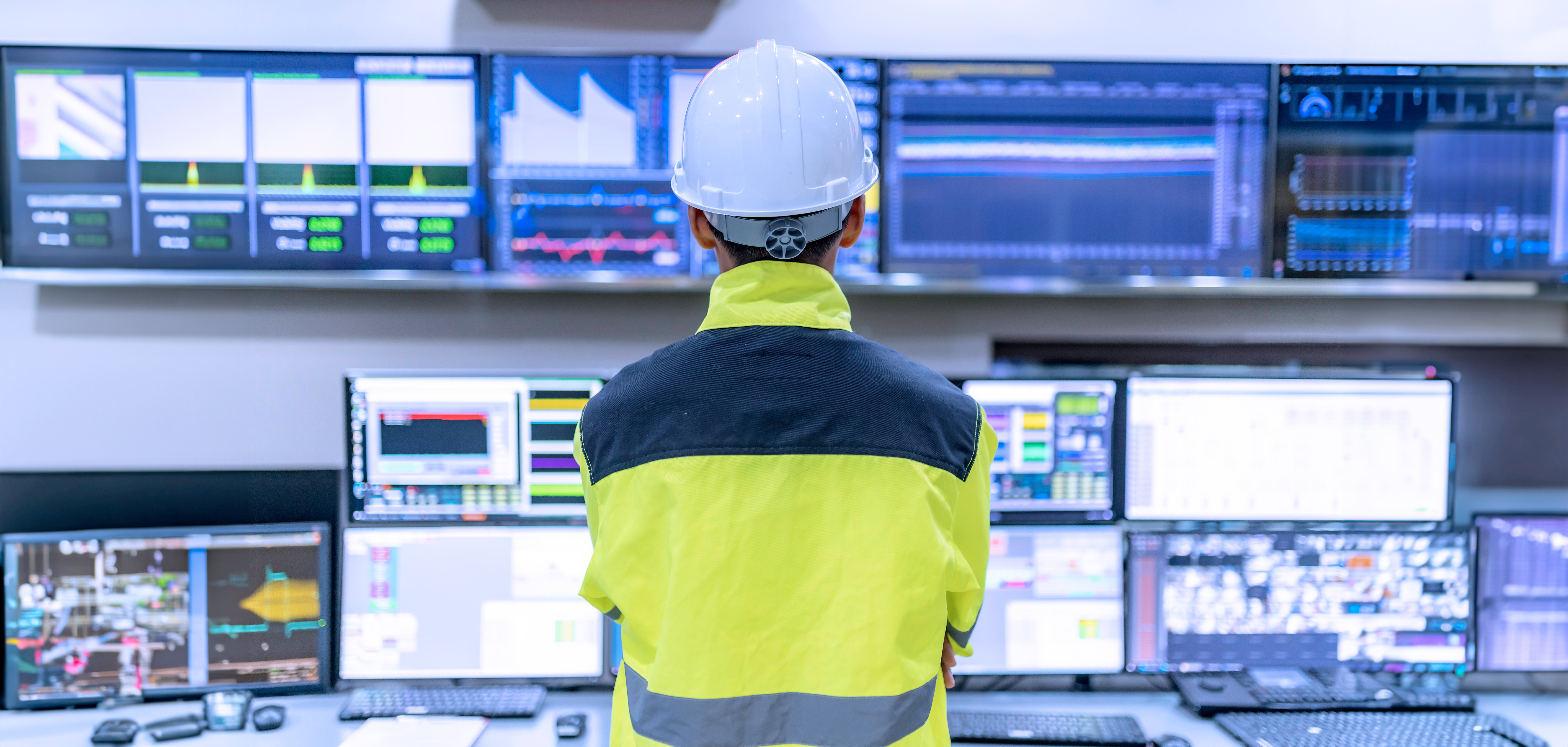 Worker in hard hat and safety vest monitoring multiple screens in control room